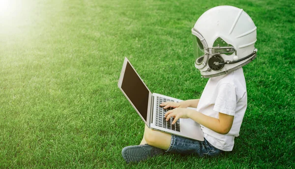 A child sits on the grass in an astronaut\'s helmet and studies on a laptop online enjoying nature
