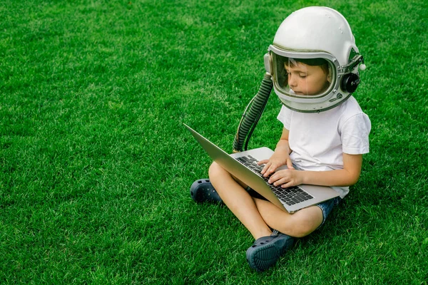 A child sits on the grass in an astronaut\'s helmet and studies on a laptop online enjoying nature