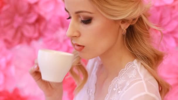 Joven hermosa novia bebiendo de taza blanca sobre fondo de flores rosadas — Vídeos de Stock