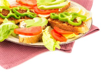 Peynirli sandviçler, kurutulmuş domatesler, biber, avokado ve tohumlar, marul, bej tabakta limon, beyaz arka planda havlu..