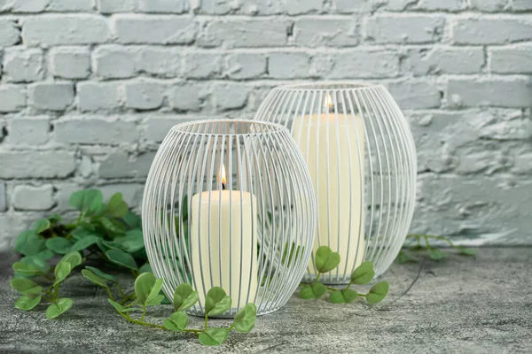 İki beyaz metal mum tutacağında iki mum ve gri tuğla duvar arka planında yapay yapraklar..