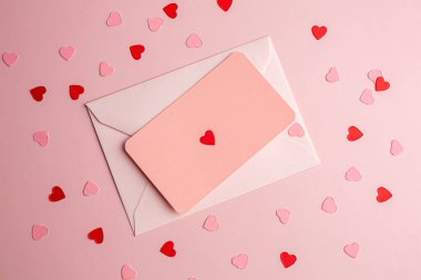 Kağıt kartlı beyaz zarf ve pembe arka planda küçük pembe ve kırmızı kalpler..