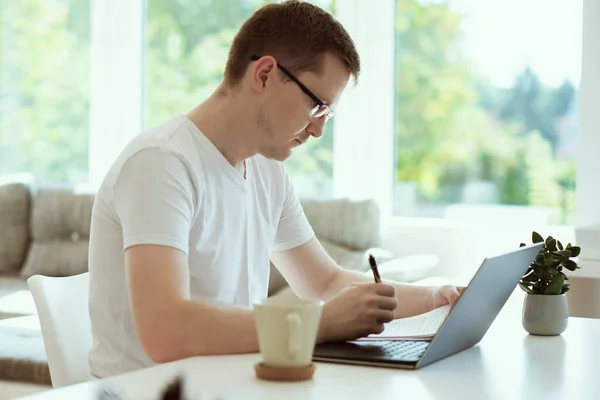严肃的年轻人在家里带着笔记本电脑上网 拿着纸坐在写字台上工作的画像 — 图库照片