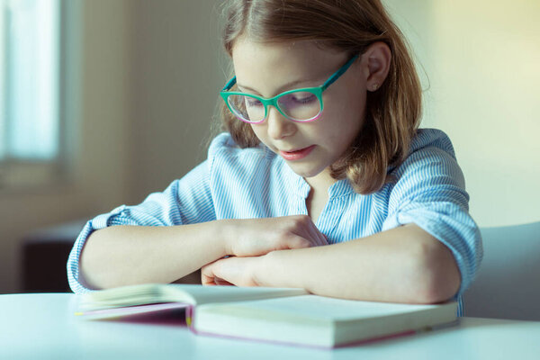 Happy pretty teen girl reading book at desk at home during pandemic distance learning 