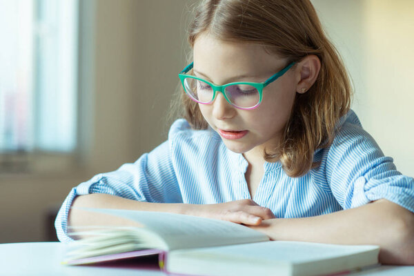 Happy pretty teen girl reading book at desk at home during pandemic distance learning 