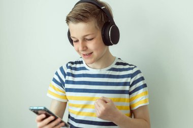 Mutlu genç çocuk beyaz arka planda kulaklık ve akıllı fonla şarkı söyleyip dans ediyor.