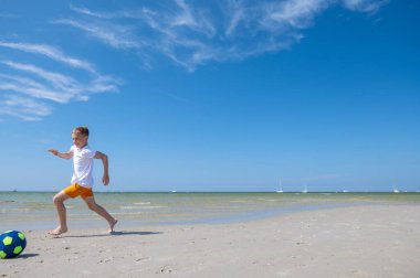Mutlu genç çocuk yaz güneşli bir günde, arka planda mavi gökyüzü ile sahilde top oynuyor.