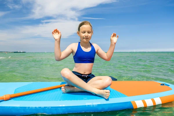 幸せな女の子座っていますオンSupパドルボード作りヨガ瞑想で澄んだ海とともに青空上の背景 — ストック写真