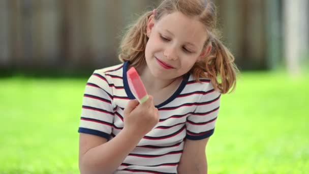 幸せな小さなティーン女の子なめるリフレッシュアイスと喜び晴れ夏時間で緑の家の裏庭 — ストック動画