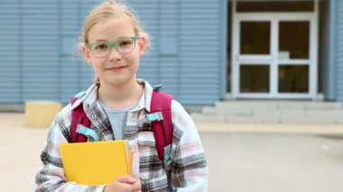 Güzel sarışın liseli kızın portresi elinde ders kitabı ve sırt çantasıyla okul çantasının önünde duruyor. 4K 60fps video