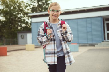 Gözlüklü, sırt çantalı, poz veren ve okulun önünde gülümseyen küçük bir kızın portresi.