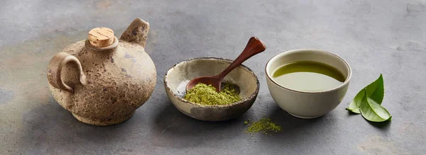 Green matcha powder with leaves and clay tea pot on graphite background