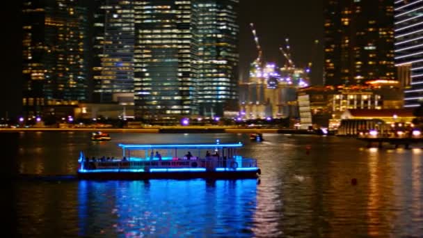 Un barco turístico. iluminado en azul. cruceros a través de Marina Bay. con el horizonte dramáticamente iluminado del distrito comercial del centro de Singapur en el fondo . — Vídeos de Stock