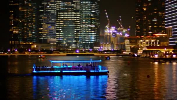 Bateau d'excursion. illuminé en bleu. croisières à travers Marina Bay. avec l'horizon dramatiquement éclairé du quartier commercial du centre-ville de Singapour en arrière-plan . — Video