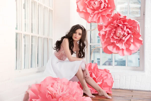 Beautiful sexy girl in a long dress with a huge pink flowers sitting by the window — Stock Photo, Image