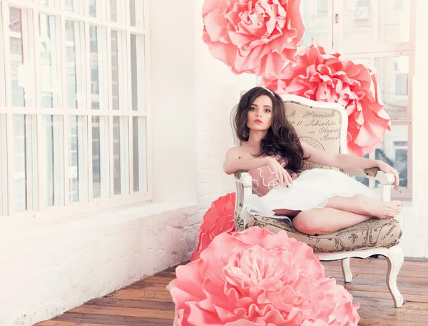 Beautiful sexy girl in a long dress with a huge pink flowers sitting by the window — Stock Photo, Image