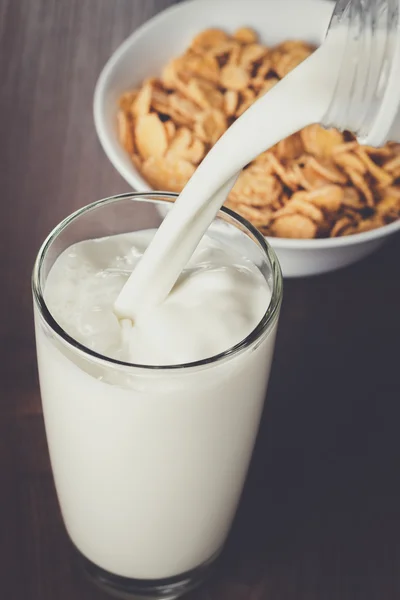 Milk pouring into glass and bowl with cornflakes — Stock Photo, Image