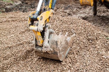 İnşaat alanında çalışan kazıcıyı kapat, kazıcı kova seviyeleri bina temelinde çakıl. Kazı kovası, gri ezilmiş taşın arka planına yakın.