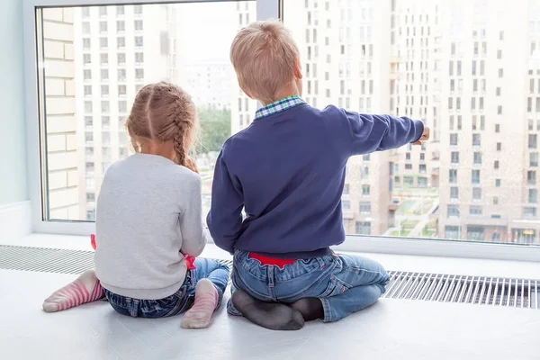 two kids sit on floor by window and admire view during quarantine. girl with pigtails and boy pointing at something outside window