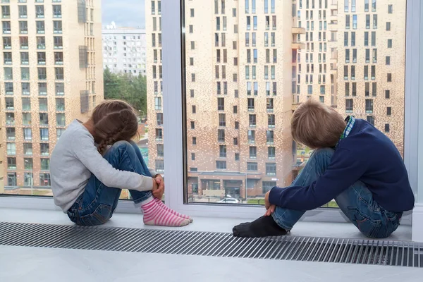 children sit on floor by window and cannot go outside during quarantine. little blond boy and girl have their hands clasped around their knees and their heads bowed and are looking out of window