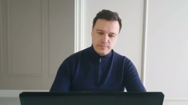 Modern ev ofisinde çalışan genç bir adam, masada oturuyor, dizüstü bilgisayar kullanıyor, görüntülü konuşma yapıyor.