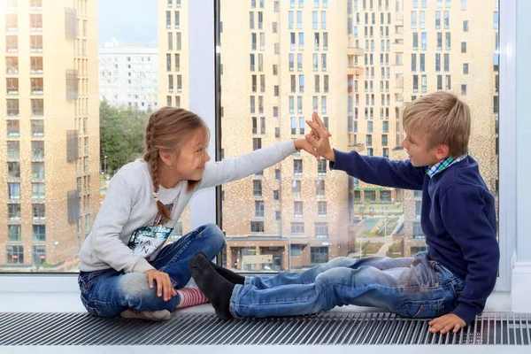 Girl gives five to boy. children sit on floor by window in new house during quarantine. Photo of children leisure house. Brother and sister are having fun at window. High five! selective focus