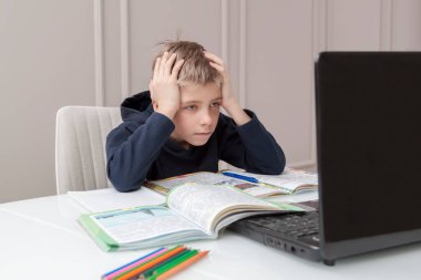 Çalışmaktan yorgun düşmüş çocuk kafasını eline koydu ve dizüstü bilgisayar ekranına baktı. Akıllı küçük çocuk ev ödevini oturma odasında yapıyor. COVID-19 salgını çocukları çevrimiçi öğrenmeye zorluyor