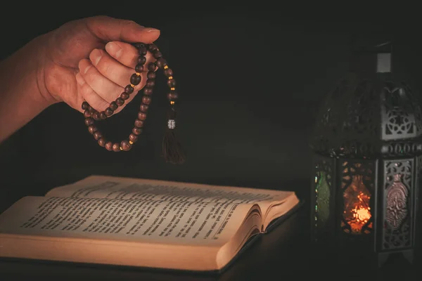 暗い背景にコーランの聖なるイスラム教徒の本の上に祈りのビーズで手 ラマダーン カリームの聖月のイスラム教徒の祝日は 輝く提灯で暗い背景にあります — ストック写真
