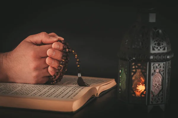 Hands with prayer beads over holy Muslim book of Koran on dark background. Muslim holiday of the holy month of Ramadan Karim on a dark background with a shining lantern