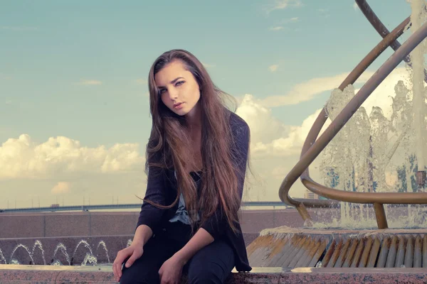 Giovane donna che indossa un abito nero è seduto vicino a una fontana in città e guardando la fotocamera, street fashion look concept . — Foto Stock