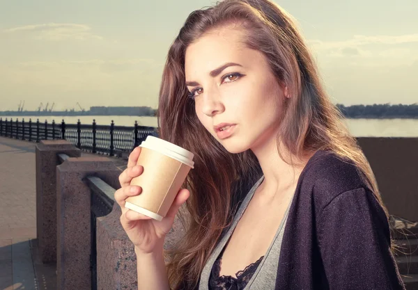 Pensive young trendy woman drinking take away coffee and standing leaning back granite fence urban scene. — Stock Photo, Image