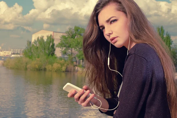 Young woman waiting your call. Dressing in a black wear,  a young caucasian lady is standing by river, holding a mobile phone, with headphones in her ears listening to the music, lost in thought. — Stockfoto