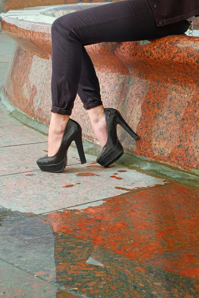 Close-up de pernas de mulher jovem na rua de paralelepípedos de granito vermelho com poça e reflexão. Pernas femininas bonitas em sapatos de moda com saltos altos no pavimento de granito — Fotografia de Stock