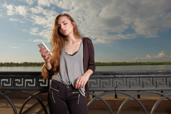 Dość młoda kobieta nachylając się powrót przeciwko balustrady na nasyp podczas korzystania jej Smartphone czytanie Sms. — Zdjęcie stockowe