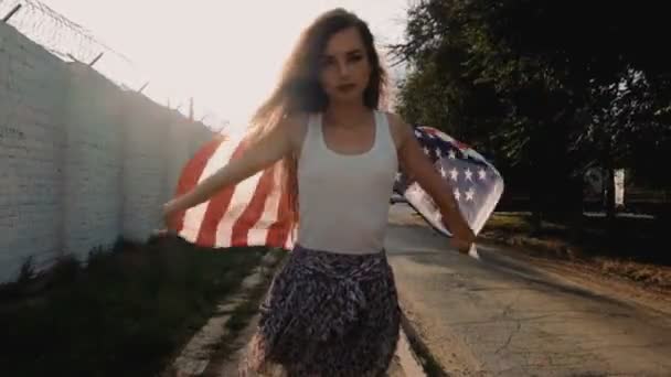 Sonriente chica americana joven con estrellas americanas y rayas bandera tejiendo en el viento, tiro de color retro. Mujer joven posando cerca de la pared con alambre de púas con bandera de EE.UU. . — Vídeos de Stock