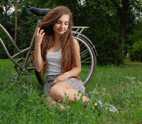 Beautiful smiling girl sitting next to bike on fresh grass with her eyes half closed — Stock Photo, Image