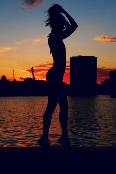 Ritratto di giovane donna come silhouette vicino al fiume. Slim ragazza sportiva è in posa in argine fiume di fronte al tramonto rosso — Foto Stock