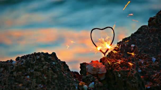 Hart-vormige sparkler op de rotsachtige kust tegenover traag bewegende zee water met warme zonsondergang lichte vlekken op het oppervlak — Stockvideo