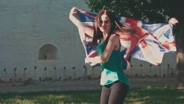 Щаслива молода жінка, яка тримає прапор Великобританії в повітрі і танцює повільно, розважаючись з державним символом — стокове відео