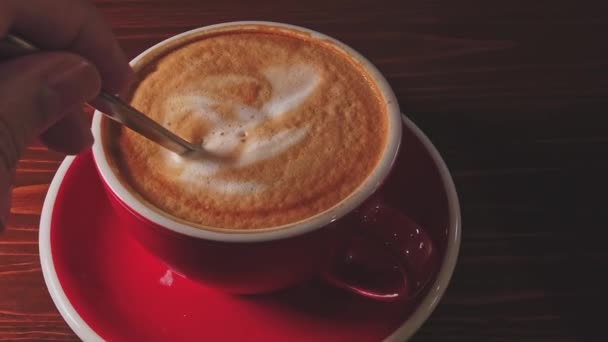 A man mixes coffee in a red cup with a spoon. On coffee thick milk foam — Stock Video