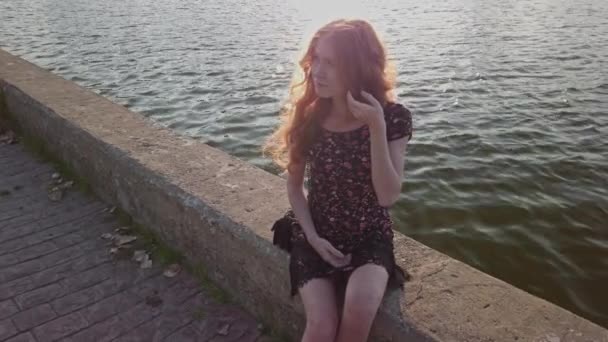 Sorgsen rödhårig flicka sitter på betong på flodsidan och tittar bort kamera, dolly i skott — Stockvideo
