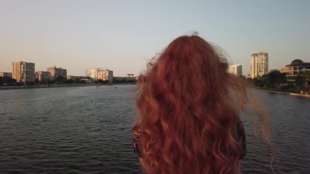 Signora pronta a guardare il fiume dal ponte, vista posteriore. Bella ragazza dai capelli rossi con lunghi capelli ondulati dalla schiena dolly in. — Video Stock