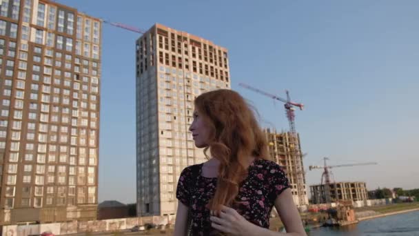 Roodharige vrouw poseren in de voorkant van hoge flatgebouwen aan de rivier. — Stockvideo