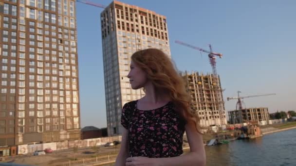 Senhora de cabelo vermelho posando na frente do canteiro de obras no lado do rio — Vídeo de Stock