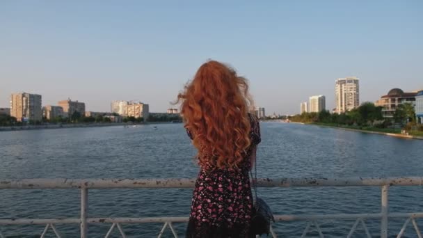 여름 옷을 입은 붉은 머리 소녀는 러시아 아스트라한의 강 위에서 바라보고 있는 다리 위를 바라본다 — 비디오