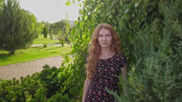 Porträt einer jungen Frau mit langen roten Haaren und Sommersprossen, die im Sommer im Park glücklich in die Kamera blickt. Konzept der Mutter Natur — Stockvideo