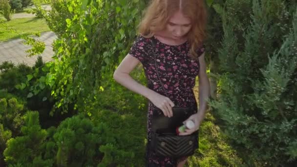 Ung rödhårig kvinna lägger en flaska i handväskan i parken — Stockvideo