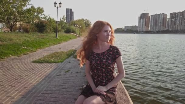 Рыжая леди в коротком летнем платье сидит на выветриваемом бетонном парапете на берегу реки со строительной стороны на заднем плане, она выглядит расслабленной и полной обещаний. — стоковое видео