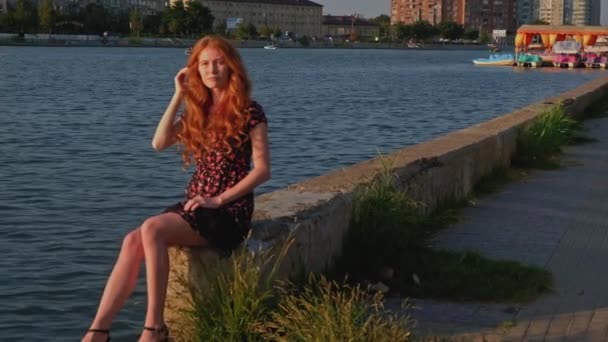 Kızıl saçlı kadın nehir kenarında betonun üzerinde oturuyor. — Stok video