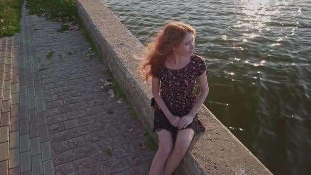 Bella signora rossa alla moda siede sul parapetto del fiume. Bellezza romantico ritratto di ragazza dai capelli rossi in abito modello floreale sul lago — Video Stock